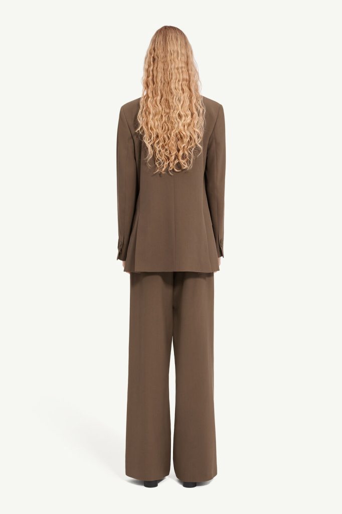 Person with long curly blond hair wearing a brown blazer and wide-leg pants, viewed from the back.