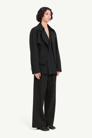 Person wearing an oversized black suit: long blazer and wide-leg trousers, black shoes, neutral pose against a light background, fashion look.