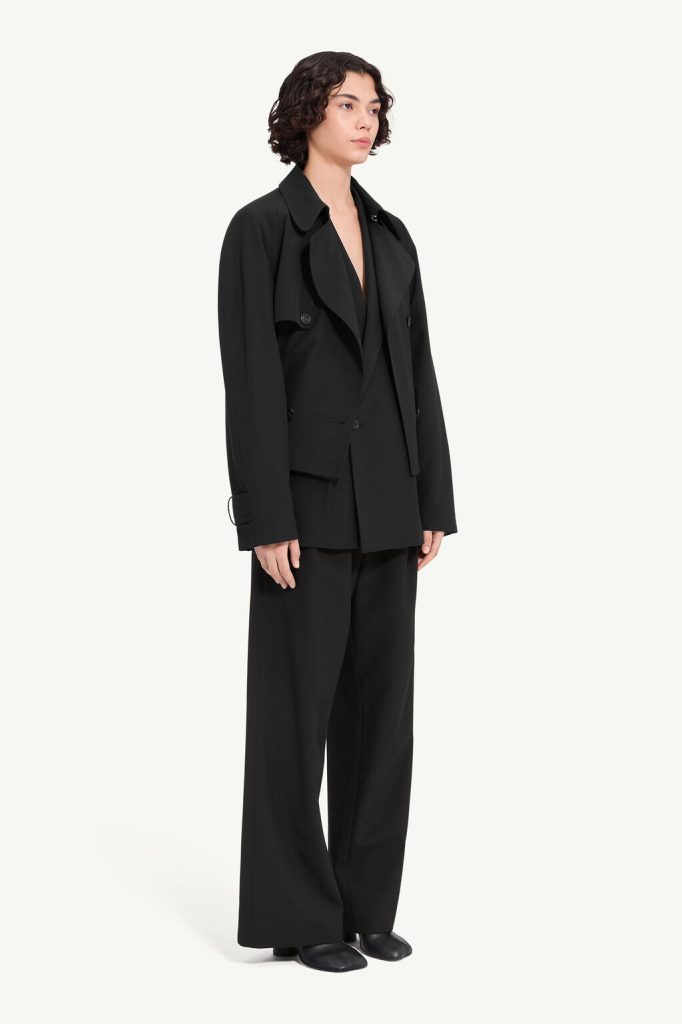 Person wearing an oversized black suit: long blazer and wide-leg trousers, black shoes, neutral pose against a light background, fashion look.