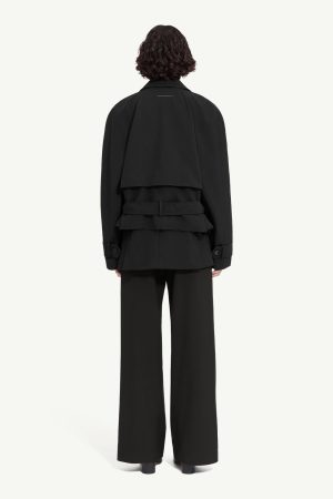 Back view of a person wearing a black oversized trench-style coat with layered back flaps, a wide belt, and wide-leg black trousers.
