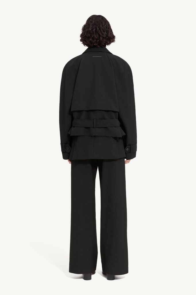 Back view of a person wearing a black oversized trench-style coat with layered back flaps, a wide belt, and wide-leg black trousers.