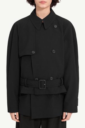 Person wearing a black double-breasted trench coat with a belt and high collar, looking straight ahead