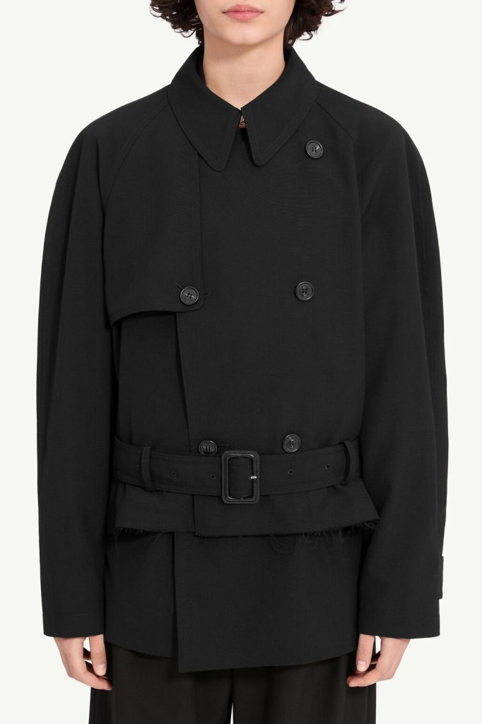 Person wearing a black double-breasted trench coat with a belt and high collar, looking straight ahead
