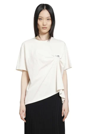 Front view of a woman wearing a loose white top with a twist detail and small logo, paired with a black pleated skirt.