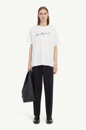 Model wearing an oversized white T-shirt with a black handwritten logo, black trousers, black boots, and holding a large black bag.