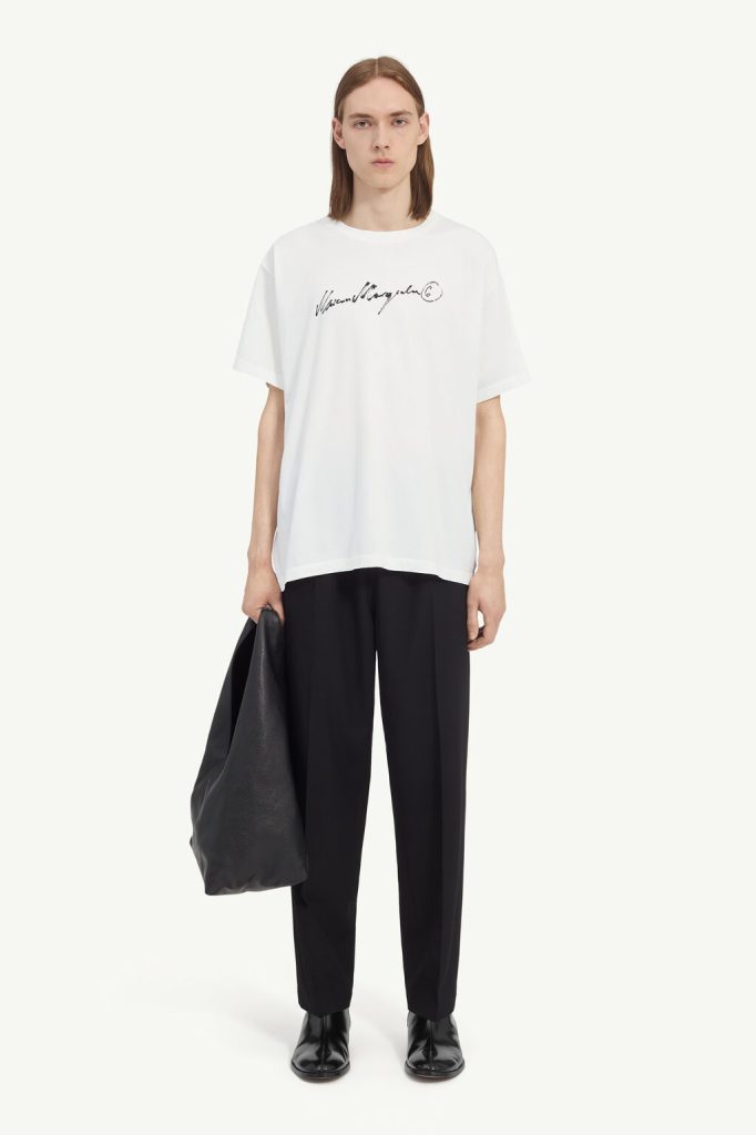 Model wearing an oversized white T-shirt with a black handwritten logo, black trousers, black boots, and holding a large black bag.