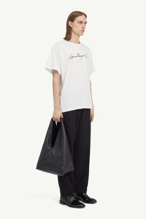 Male model in an oversized white T-shirt with black cursive logo, wide-leg black trousers, black loafers, and a large black tote bag on his left side.