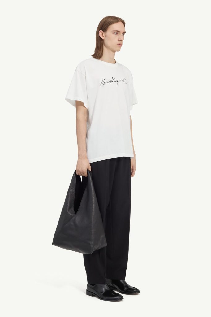 Male model in an oversized white T-shirt with black cursive logo, wide-leg black trousers, black loafers, and a large black tote bag on his left side.