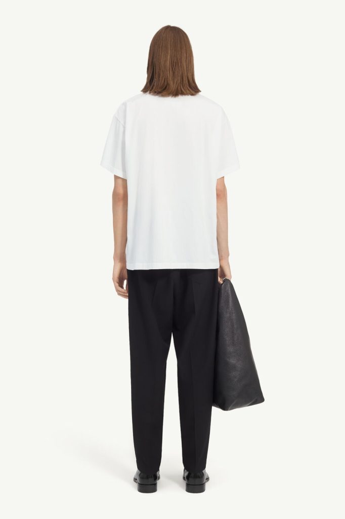 Back view of a person wearing a white oversized T-shirt, black trousers, and black shoes, holding a large black tote bag at their side.