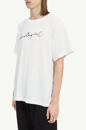 White oversized T-shirt with a black handwritten logo on the chest, worn by a person photographed from the side.