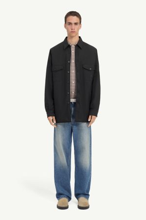 Male model wearing a black button-front jacket over a plaid shirt with loose blue jeans and tan shoes against a light background.