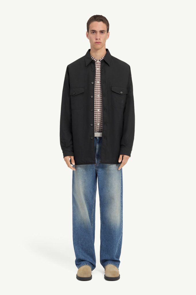 Male model wearing a black button-front jacket over a plaid shirt with loose blue jeans and tan shoes against a light background.