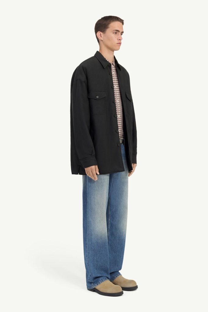 Male model in a black utility jacket over a checkered shirt, paired with wide-leg blue jeans and tan shoes, shown in profile.