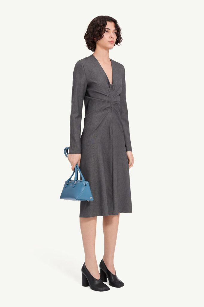 Woman in a knee-length grey V-neck dress, black chunky-heel shoes, carrying a small blue handbag.