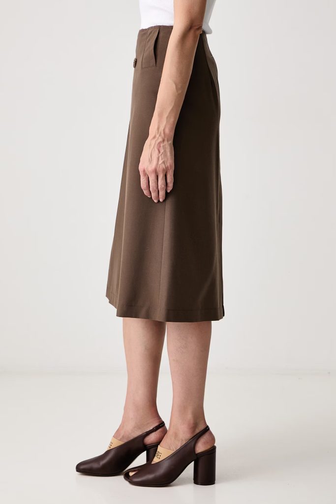 Side view of a person wearing a brown midi skirt with a pleated waist detail, paired with brown block-heeled sandals and a white top.