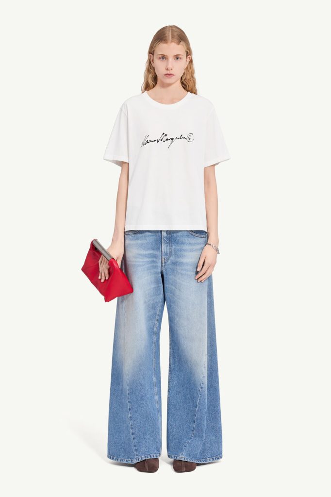 Model in a white oversized logo tee and wide-leg blue jeans, holding a red clutch bag with brown shoes.