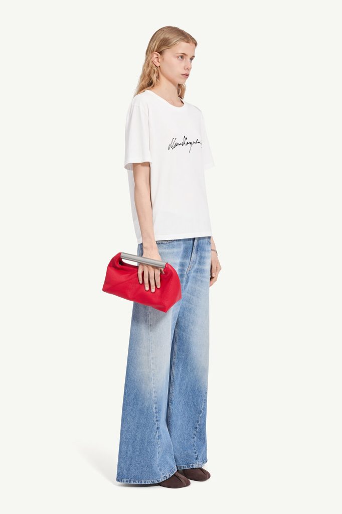 Model in a white oversized T-shirt with black script logo, light-wash wide-leg jeans, brown flats, and a red clutch with a silver handle they hold at the side.