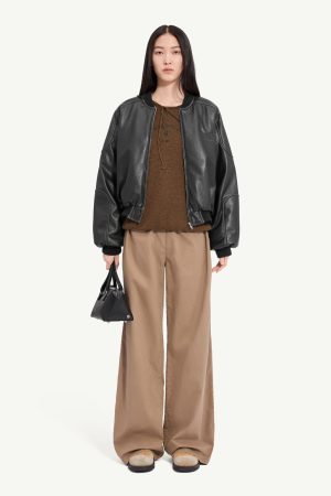 Full-body portrait of a woman wearing a black leather bomber jacket, brown knit top, tan wide-leg pants, and a small black handbag.