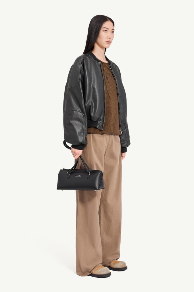 Model wearing a black leather bomber jacket over a brown knit top with wide tan trousers, carrying a small black handbag.