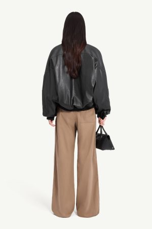 Back view of a person with long dark hair wearing a glossy black oversized bomber jacket and tan wide-leg pants, holding a small black handbag.