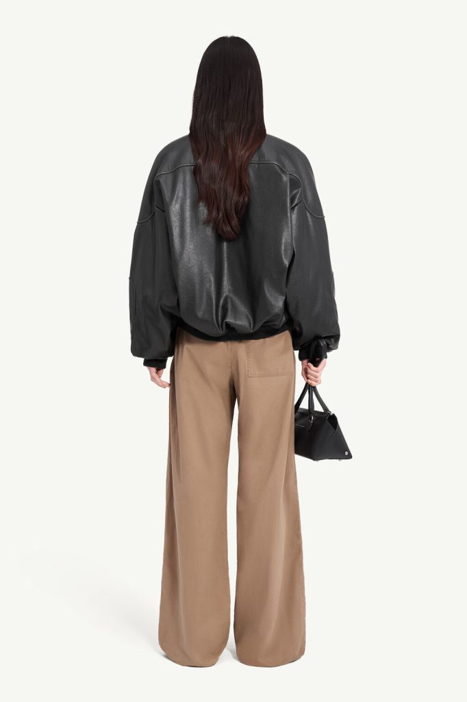 Back view of a person with long dark hair wearing a glossy black oversized bomber jacket and tan wide-leg pants, holding a small black handbag.
