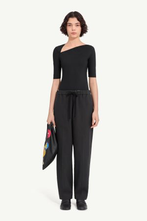 Model wearing a black, one-shoulder top with short sleeves and black wide-leg pants, holding a black pouch with colorful patches.