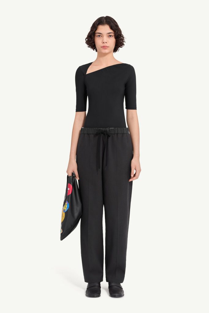 Model wearing a black, one-shoulder top with short sleeves and black wide-leg pants, holding a black pouch with colorful patches.
