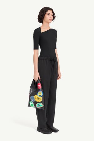 Woman in an all-black outfit: fitted one-shoulder top with short sleeves and wide-leg pants, holding a black tote covered in colorful patches.