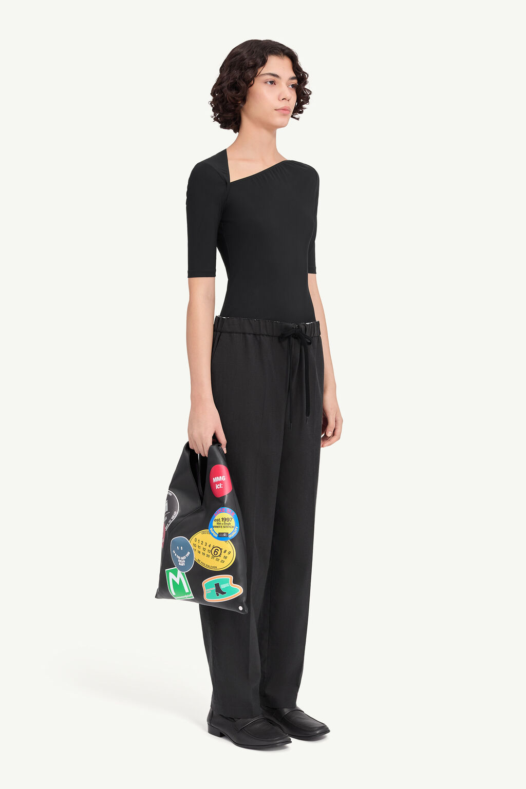 IMG_2971 - New Trend Woman in an all-black outfit: fitted one-shoulder top with short sleeves and wide-leg pants, holding a black tote covered in colorful patches.