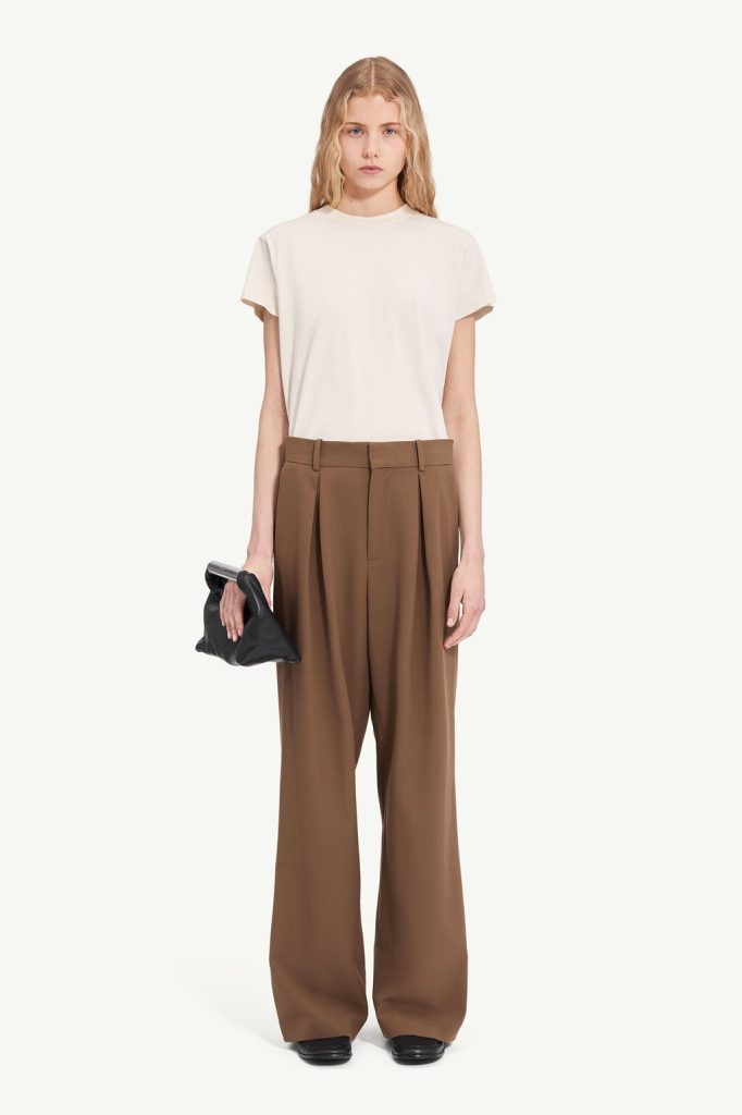 Full-body shot of a woman in a cream T-shirt and brown wide-leg trousers, holding a black clutch.