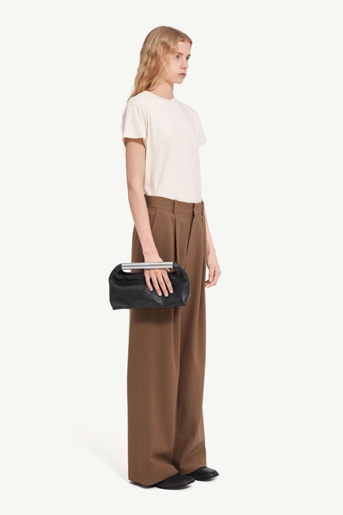 Model in a cream T-shirt and brown high-waisted wide-leg trousers, holding a black handbag with a silver bar handle.