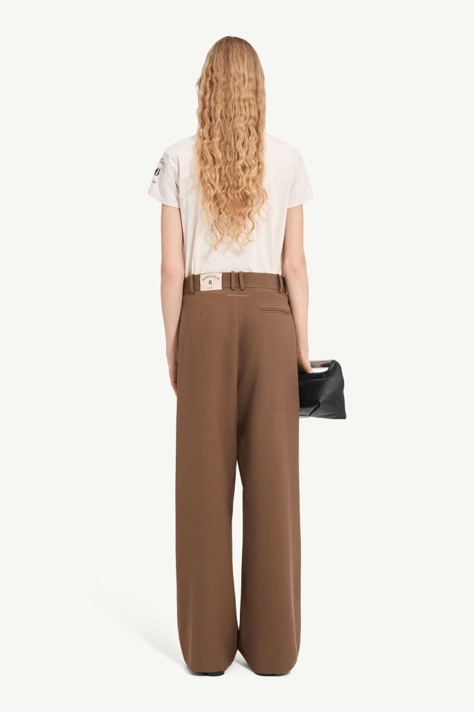 Woman with long wavy blonde hair, wearing a white T-shirt and brown wide-leg pants, viewed from behind while holding a black clutch.