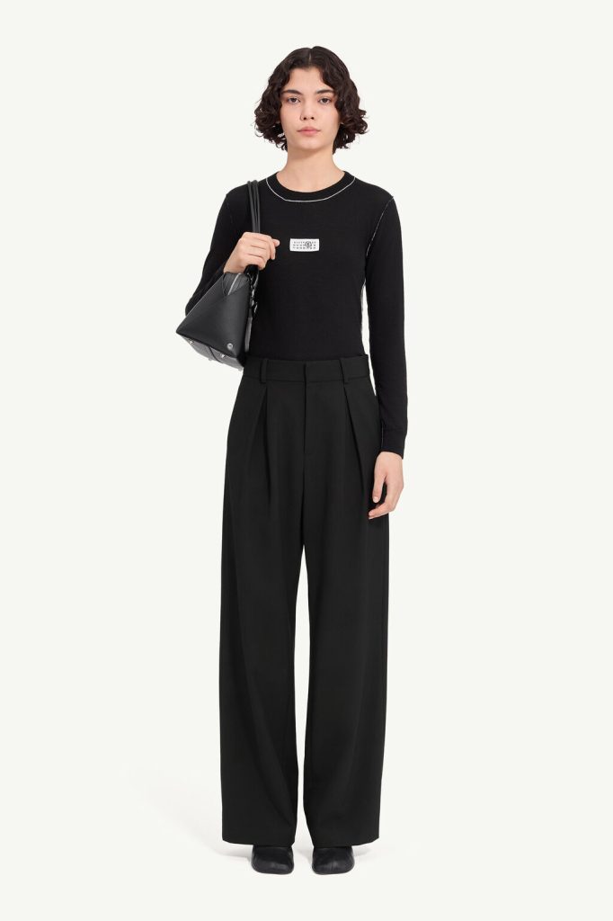 Woman in a black long-sleeve top with white stitching and wide-leg black trousers, carrying a gray shoulder bag.