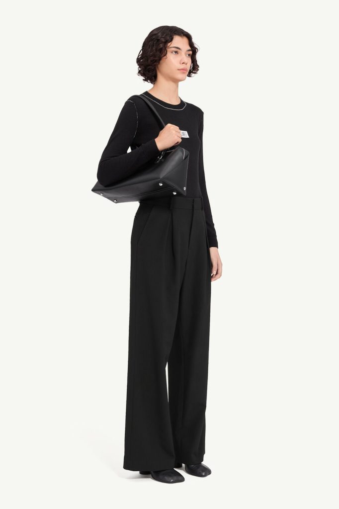 Female model in all-black outfit (long-sleeve top and wide-leg trousers) carrying a black leather shoulder bag over her right shoulder against a pale backdrop.