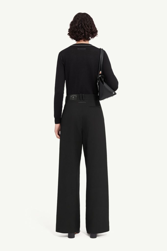 Back view of a person wearing a black long-sleeve top and black wide-leg pants, with a black shoulder bag resting on the left side.