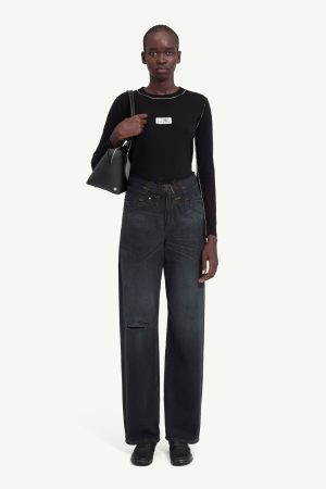 Full-body shot of a model wearing a black long-sleeve top, wide dark jeans with a knee rip, black shoes, and a black shoulder bag against a light background.