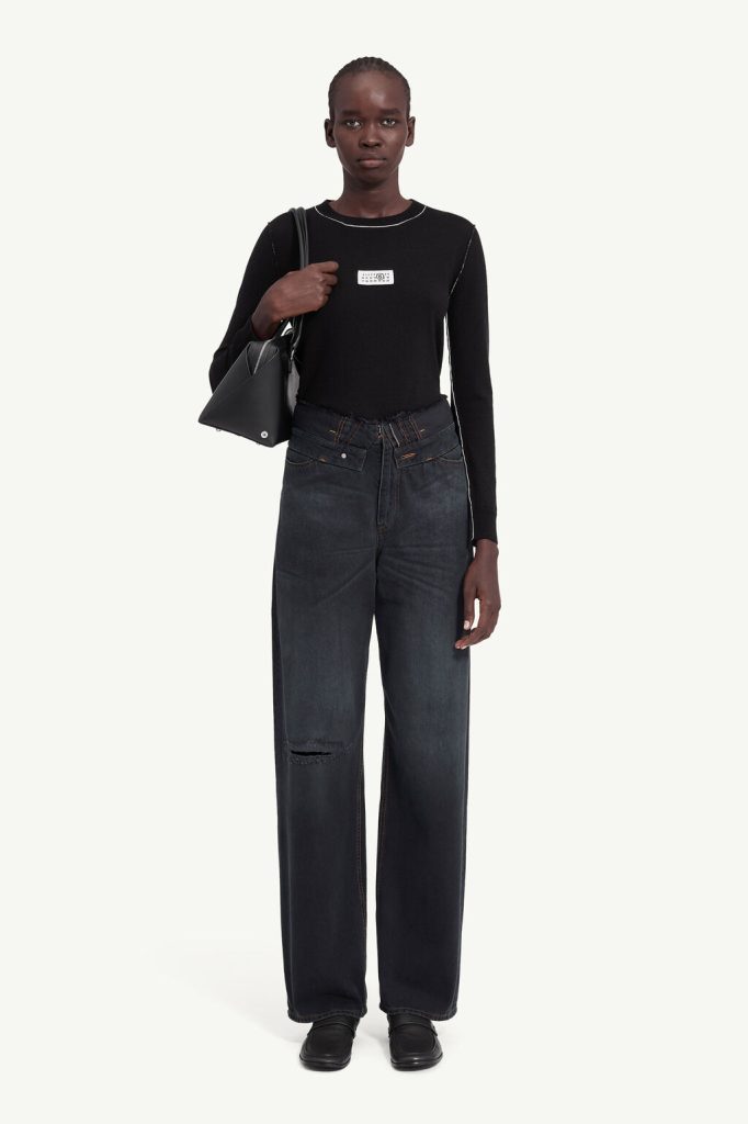 Full-body shot of a model wearing a black long-sleeve top, wide dark jeans with a knee rip, black shoes, and a black shoulder bag against a light background.