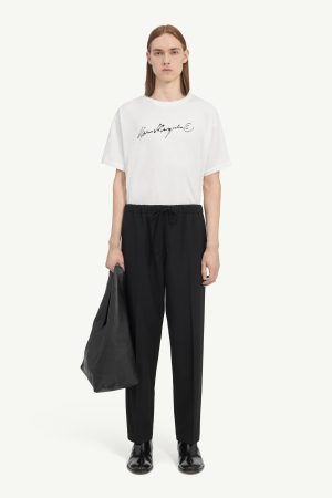 Full-body portrait of a person wearing a white logo T-shirt, black drawstring pants, black ankle boots, and a black bag held at their side.