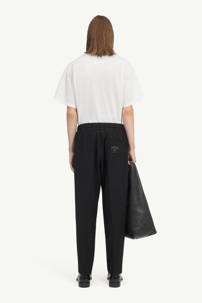 Back view of person in a white T-shirt, black elastic-waist pants, black shoes, carrying a large black tote.