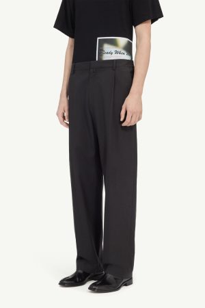Person standing in a black T-shirt and dark wide-leg trousers; a rectangular graphic patch is visible at the back of the shirt near the waist.