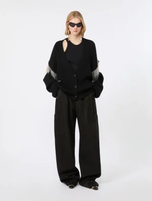 Model in an oversized black cardigan with fringe sleeves, black top and wide cargo pants with a sleek sunglasses look.