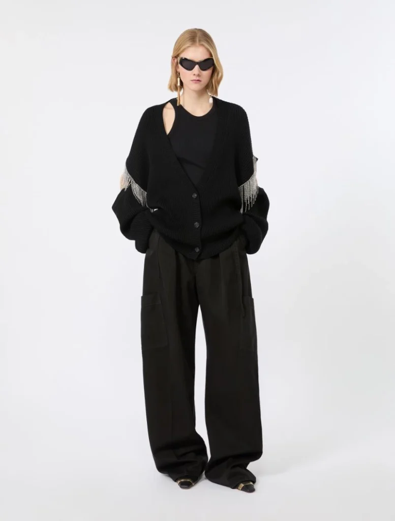 Model in an oversized black cardigan with fringe sleeves, black top and wide cargo pants with a sleek sunglasses look.
