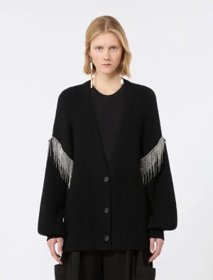 Model wearing a black chunky cardigan with silver fringe on the sleeves, standing against a light background.