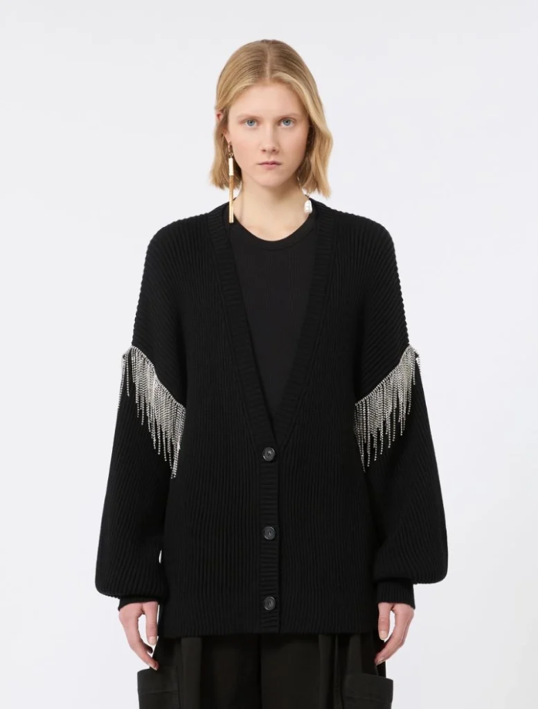 Model wearing a black chunky cardigan with silver fringe on the sleeves, standing against a light background.