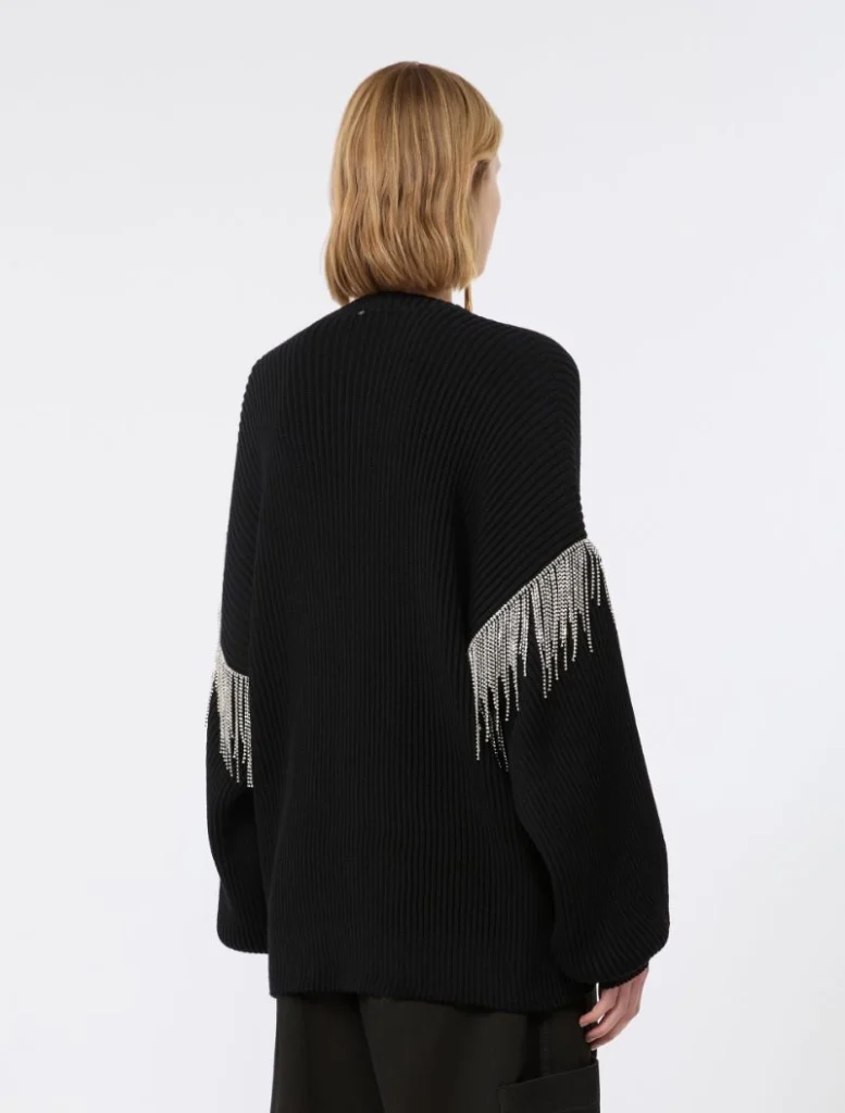 Back view of a person wearing an oversized black knit sweater with beaded fringe along the sleeves, blonde hair visible from behind.