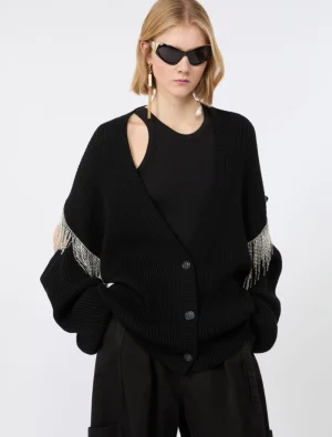 Fashion model wearing a black oversized knit cardigan with beaded fringe on the sleeves, paired with a black top and dark sunglasses.