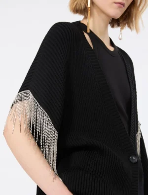 Woman wearing a black ribbed cardigan with metallic fringe streaming from the sleeves, over a black top with gold earrings visible at the ear
