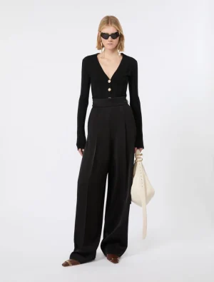 Model in a black V-neck cardigan with gold buttons and high‑waisted wide‑leg pants, holding a cream handbag and wearing sunglasses.