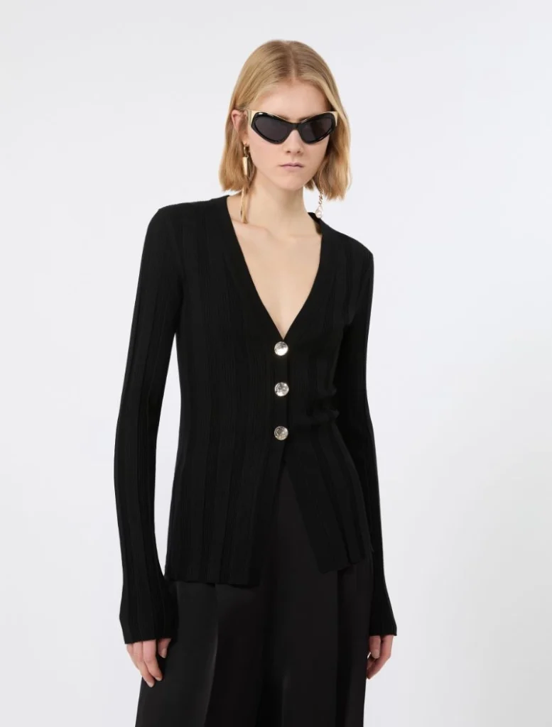 Model wearing a black ribbed cardigan with large silver buttons over a deep V-neck, paired with black pants and sunglasses.