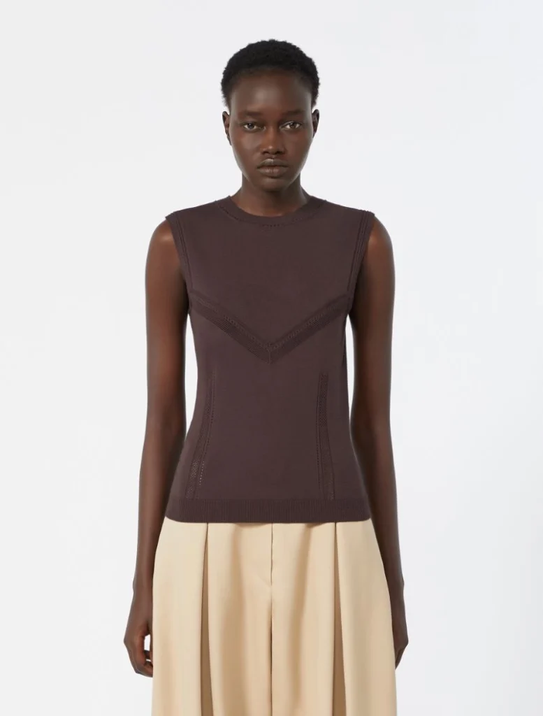 Female model wearing a sleeveless dark brown knit top with chevron V-stitch details, paired with a beige skirt, standing against a light background.